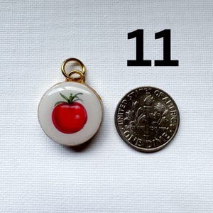 May include: A round pendant featuring a red tomato illustration on a white background with a gold-toned metal jump ring. The pendant is positioned next to a United States dime for scale, with the number 11 printed in black above the items.