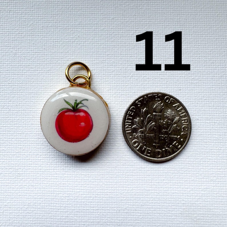May include: A round pendant featuring a red tomato illustration on a white background with a gold-toned metal jump ring. The pendant is positioned next to a United States dime for scale, with the number 11 printed in black above the items.