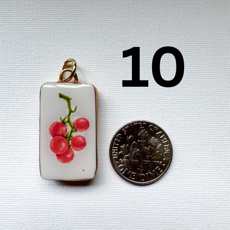 May include: A rectangular white pendant featuring a graphic of a cluster of red cherry tomatoes on a green vine, topped with a gold-toned metal bail. The pendant is positioned next to a United States dime for scale, with the number 10 above.