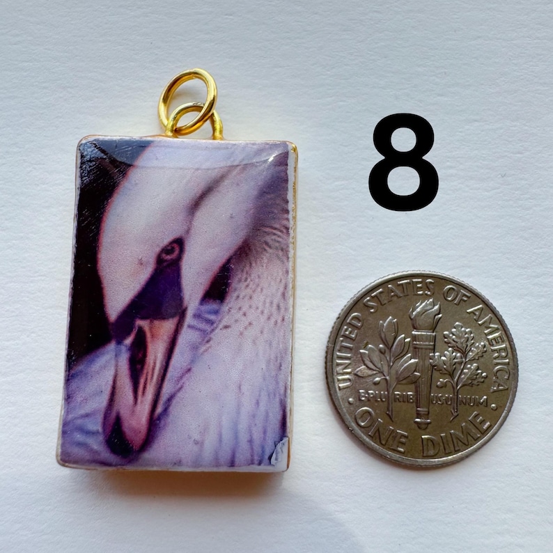 May include: A rectangular pendant with a close-up image of a swan's head, featuring white feathers and a dark beak. The pendant has a gold-colored bail. A dime is next to the pendant for size comparison. The number 8 is in the background.