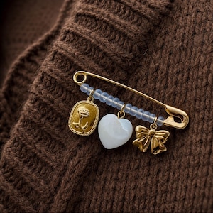 May include: A gold-tone safety pin brooch featuring a row of small, translucent light blue beads. Three charms hang from the pin: a gold oval pendant with a rose relief, a smooth white heart-shaped charm, and a gold-tone bow-shaped charm.