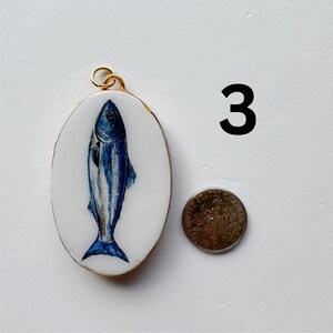 May include: An oval pendant featuring a blue fish illustration on a white background with a gold-toned metal rim and jump ring. The number 3 is printed in black to the right, next to a US dime for scale, which is 0.705 inches in diameter.