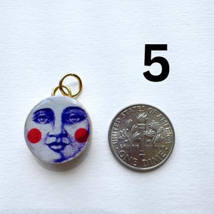 May include: A round pendant with a smiling moon face design, featuring blue facial details and red cheeks, set in a gold-colored frame. Next to it is a US dime coin for size comparison. The number "5" is in the background.