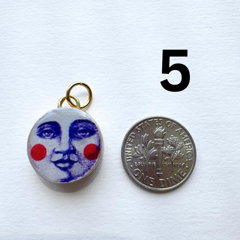 May include: A round pendant with a smiling moon face design, featuring blue facial details and red cheeks, set in a gold-colored frame. Next to it is a US dime coin for size comparison. The number "5" is in the background.