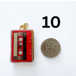 K&ouml;nnte beinhalten: Ein roter Kassetten-Anh&auml;nger mit einem goldenen Biegering, der die Aufschrift C-60 und B auf dem Etikett tr&auml;gt. Der Anh&auml;nger ist neben einer amerikanischen 10-Cent-M&uuml;nze als Gr&ouml;&szlig;envergleich abgebildet, mit der Zahl 10 in Schwarz oben rechts.