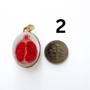 K&ouml;nnte beinhalten: Ein ovaler Anh&auml;nger mit einer gemalten Illustration eines halbierten Granatapfels mit leuchtend roten Kernen auf wei&szlig;em Grund, eingefasst in goldfarbenes Metall mit einem Biegering, neben einem US-Dime zur Gr&ouml;&szlig;enorientierung, gekennzeichnet mit der Zahl 2.