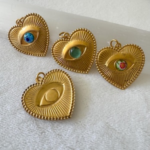 May include: Four gold-colored heart-shaped pendants with an eye design. Each pendant has a different colored eye, including blue, green, and a floral pattern. The pendants have a beaded border and a loop for hanging.