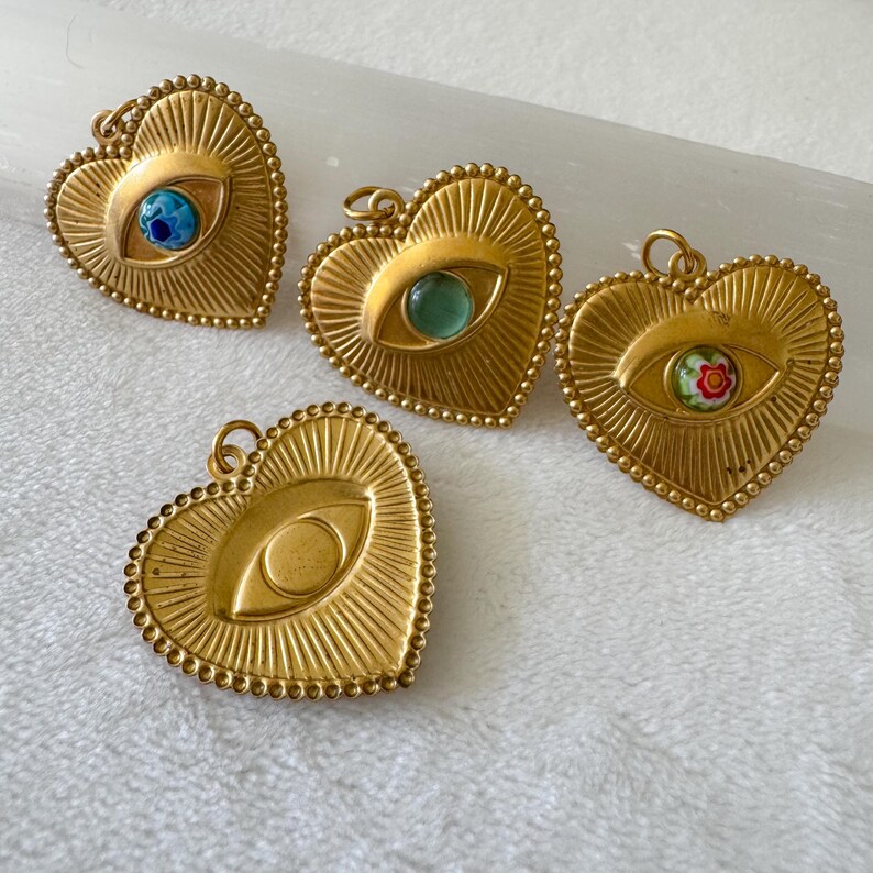 May include: Four gold-colored heart-shaped pendants with an eye design. Each pendant has a different colored eye, including blue, green, and a floral pattern. The pendants have a beaded border and a loop for hanging.