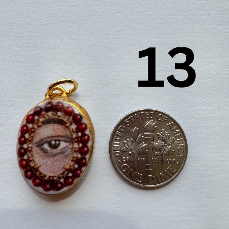 May include: An oval pendant with a painted eye design, surrounded by red beads, and a gold-colored bail. A dime is next to the pendant for size comparison. The number "13" is printed in black.