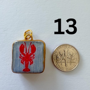May include: A square pendant with a gold-colored metal frame and a jump ring, featuring a red lobster graphic on a light blue and white striped background. A United States one dime coin is placed next to the pendant for scale, and the number 13 is printed in black above the items.