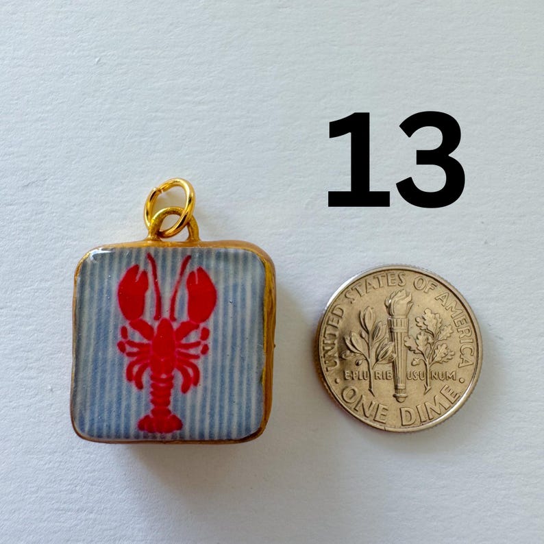 May include: A square pendant with a gold-colored metal frame and a jump ring, featuring a red lobster graphic on a light blue and white striped background. A United States one dime coin is placed next to the pendant for scale, and the number 13 is printed in black above the items.