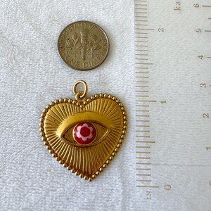 May include: A gold-colored heart-shaped pendant with an eye design and a red and white flower in the center. The pendant is approximately 1 inch in length. A U.S. dime is next to the pendant for size comparison.