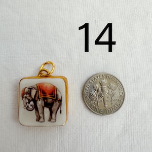 May include: Square pendant with a gold-colored frame, featuring a painted elephant with a red blanket. A dime is next to the pendant. The number "14" is in the background.