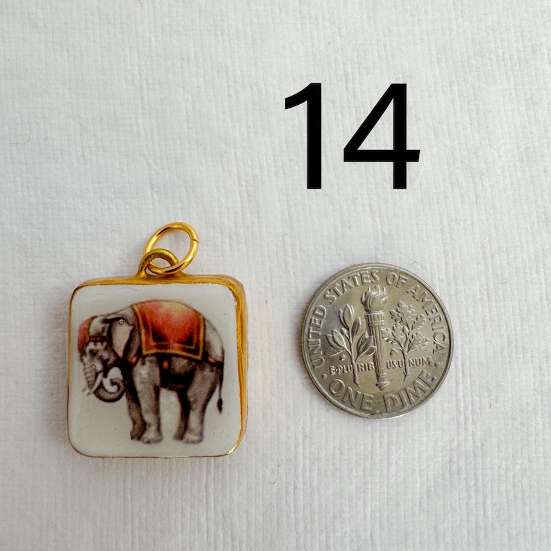 May include: Square pendant with a gold-colored frame, featuring a painted elephant with a red blanket. A dime is next to the pendant. The number "14" is in the background.