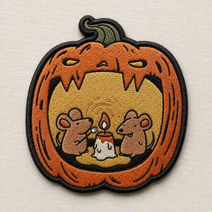May include: An embroidered patch shaped like a carved orange pumpkin with a green stem. Inside the pumpkin, two brown mice sit on a yellow background, one holding a marshmallow over a lit white candle with a small orange flame.
