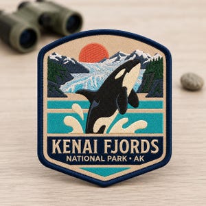 May include: An embroidered patch featuring a black and white orca breaching from turquoise water. The background shows snow-capped mountains, dark green pine trees, and a red sun. The text reads KENAI FJORDS NATIONAL PARK AK in dark blue.