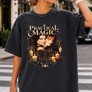May include: A charcoal grey short-sleeve t-shirt featuring a graphic print with the text Practical Magic, two women, a black cat, candles, and a book. The design includes celestial motifs like moons and stars, creating a detailed, dark-toned illustration.