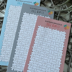 Könnte beinhalten: Drei unterschiedlich farbige Puzzle-Karten mit dem Text "KLEINGELDPUZZLE 112 EURO" auf jeder Karte. Die Puzzles bestehen aus kleinen Quadraten, in denen verschiedene Euro-Beträge gedruckt sind.