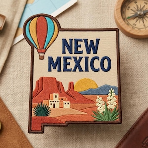 May include: An embroidered patch shaped like the state of New Mexico, featuring a colorful hot air balloon, red rock mesas, a desert landscape with adobe buildings, a yellow sun, and green yucca plants with the text NEW MEXICO in blue.