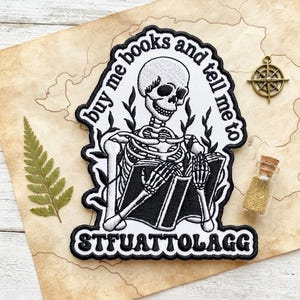May include: An embroidered patch featuring a white skeleton reading a black book, surrounded by black leafy vines. The text reads "buy me books and tell me to STFUATTOLAGG" in black lettering against a white background with a thick black border.
