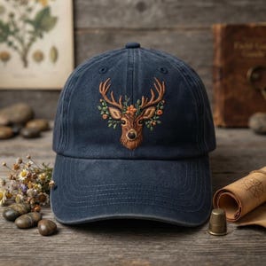 May include: A navy blue baseball cap featuring an embroidered design of a brown deer head adorned with green leaves and small orange flowers. The cap has a structured crown, a curved brim, and a button at the top centre of the fabric panels.