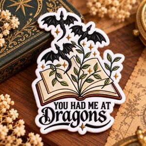 May include: An embroidered patch featuring an open book with green leafy vines growing from the pages, three black flying dragons, and small gold stars. The text "YOU HAD ME AT DRAGONS" is embroidered in black at the bottom of the white patch.