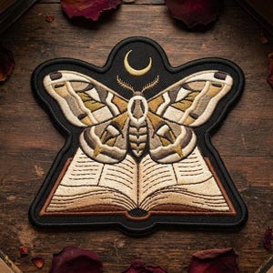 May include: An embroidered patch featuring a moth with tan, brown, and cream wings positioned above an open book with visible pages. A gold crescent moon is stitched above the moth's head. The entire design is outlined in black thread.