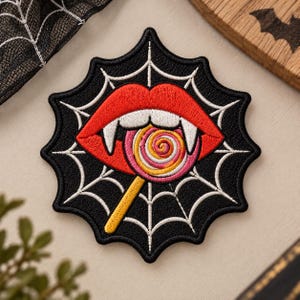 May include: A black embroidered patch shaped like a spiderweb featuring a pair of red lips with white fangs biting into a round, multi-colored swirl lollipop with a yellow stick. The design is centered on a black background with white web lines.