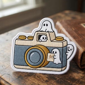 May include: An embroidered patch featuring a vintage-style camera in shades of blue, beige, and gold. Three small white ghosts are depicted: one on top, one in the viewfinder, and one holding a small square photo with a star icon on the front.