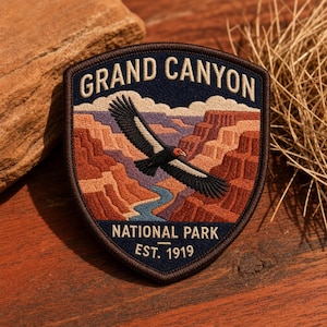 May include: A shield-shaped embroidered patch featuring a Grand Canyon landscape with a flying condor, a winding blue river, and layered red rock formations. The text reads GRAND CANYON NATIONAL PARK EST. 1919 in cream-colored thread on a dark blue background.