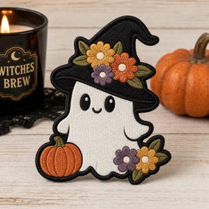 Può includere: Una toppa ricamata raffigurante un fantasma bianco che indossa un cappello da strega nero decorato con fiori gialli, arancioni e viola. Il fantasma tiene in mano una piccola zucca arancione e alla base si trova un grappolo di fiori.