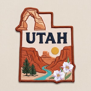 May include: An embroidered patch shaped like the state of Utah, featuring a tan background with the word UTAH in dark blue. The design includes a brown sandstone arch, red rock canyons, a winding blue river, a yellow sun, two green pine trees, and white flowers.