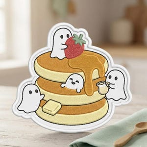 May include: An embroidered patch featuring a stack of three golden-brown pancakes topped with a red strawberry, a pat of yellow butter, and golden syrup. Three small white ghosts are interacting with the pancakes, one holding a small pitcher.