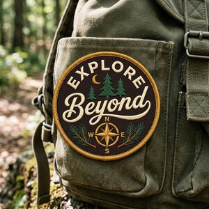 May include: A circular embroidered patch attached to an olive green canvas backpack. The patch features a dark brown background with the text "EXPLORE Beyond" in cream, a crescent moon, two green pine trees, and a gold compass with cardinal directions.