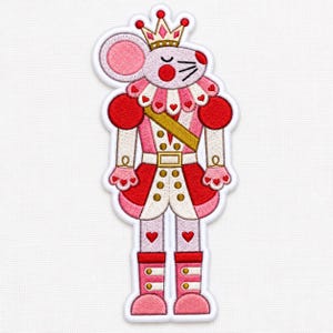 May include: An embroidered patch featuring a stylized mouse nutcracker character wearing a gold crown, a pink and white uniform with red accents, a gold sash, and heart-shaped details. The design includes a white border on a white fabric background.