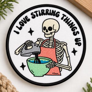 May include: A circular embroidered patch with a white background and black border. It features a skeleton wearing a coral apron, using a gray hand mixer in a light green bowl. The text I LOVE STIRRING THINGS UP is arched above with black stars.