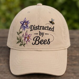 May include: A beige baseball cap featuring an embroidered design of two purple columbine flowers on the left side and a small bee on the right. The text "Distracted by Bees" is embroidered in black in the center with a dashed flight path line.