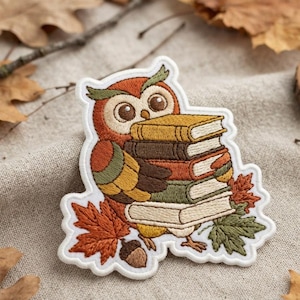 May include: An embroidered patch featuring a brown owl with large eyes holding a stack of four books in various colors. The design includes orange and green maple leaves and a small brown acorn at the base, all outlined in white thread.