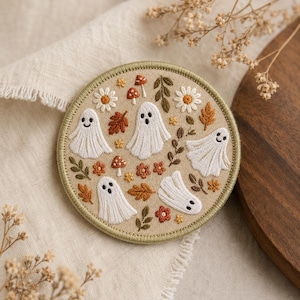 May include: A circular embroidered patch on a beige fabric base featuring four white ghosts with black eyes and mouths, surrounded by small orange and brown autumn leaves, red and white mushrooms, and white daisy-like flowers with yellow centers.