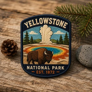 May include: An embroidered patch featuring a brown bison standing before a colorful geothermal pool and a geyser erupting against a blue sky. The text reads YELLOWSTONE NATIONAL PARK EST. 1872 in cream and orange lettering on a dark blue border.