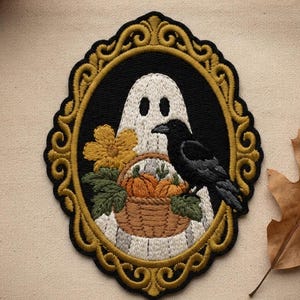 May include: An oval embroidered patch featuring a white ghost holding a brown basket filled with orange pumpkins and green leaves. A black crow sits on the basket next to a yellow flower. The design is enclosed in a decorative gold frame.
