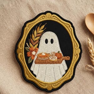 May include: An embroidered patch featuring a white ghost holding a wooden board with a pumpkin pie, cinnamon rolls, and a small pumpkin. The ghost is framed by a gold-colored ornate border against a black background, with wheat and orange flowers.