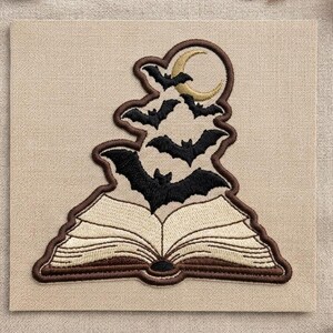 May include: An embroidered patch featuring an open book with cream-colored pages and a dark brown cover. Rising from the center of the book are several black bats flying toward a golden crescent moon, all outlined with a dark brown border.