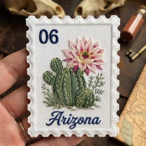 May include: A rectangular white embroidered patch shaped like a postage stamp featuring a green cactus with a large pink flower. The number 06 is stitched in dark blue at the top left, and the word Arizona is stitched in dark blue at the bottom.