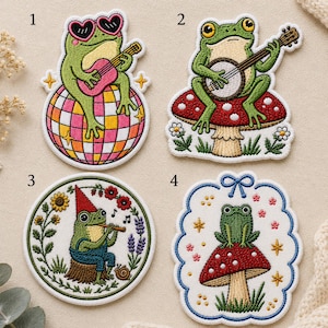 May include: Four embroidered patches featuring frogs. Patch 1 shows a frog with heart-shaped sunglasses playing a guitar on a disco ball. Patch 2 shows a frog playing a banjo on a mushroom. Patch 3 shows a gnome-hat frog playing a flute. Patch 4 shows a frog on a mushroom with a blue bow.