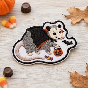 May include: An embroidered patch featuring a gray and white opossum wearing a black cape with an orange collar. The opossum holds an orange jack-o'-lantern bucket, with a small purple candy and a black bat silhouette nearby on a white background.