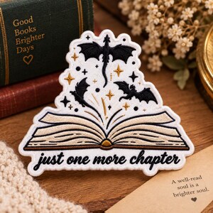 May include: A white embroidered patch featuring an open book with beige pages and black outlines. Above the book are three black dragon silhouettes and gold star accents. The text "just one more chapter" is embroidered in black at the bottom.