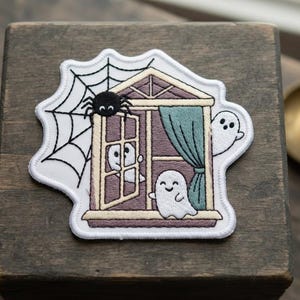May include: An embroidered patch featuring a house window with a spider and web on the left, a teal curtain, and three white ghosts. One ghost is inside the window frame, one is on the sill, and one is peeking from the right side of the house.