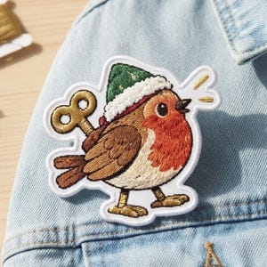 May include: An embroidered patch featuring a round robin bird wearing a green hat with a white trim. The bird has a bright orange chest, brown wings, and a gold wind-up key protruding from its back. The patch is white-bordered and stitched.