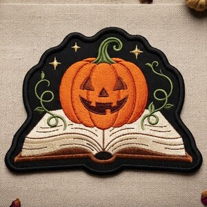 May include: An embroidered patch featuring a carved orange jack-o'-lantern pumpkin resting on an open book with cream-colored pages. The design includes green swirling vines and small yellow stars against a black background with a black border.